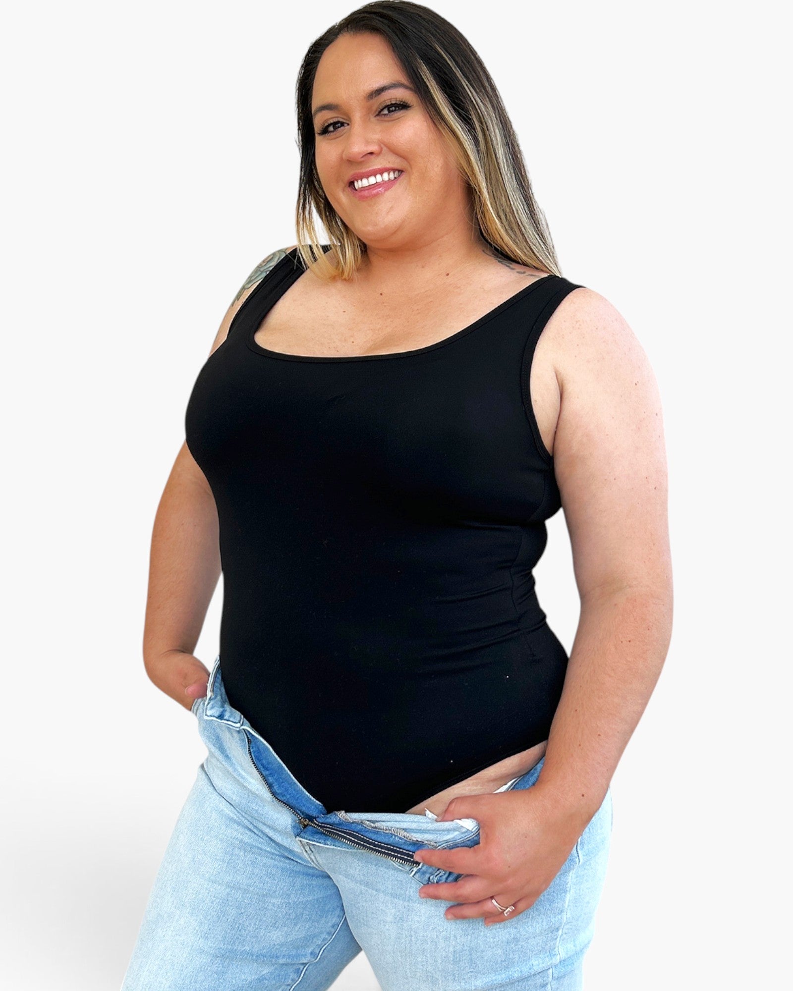 A woman wearing a black scoop-neck tank top and light-wash blue jeans, standing against a plain white background.