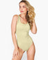 A woman modeling a sleeveless scoop-neck one-piece swimsuit in a light sage green color, set against a plain light gray studio background.