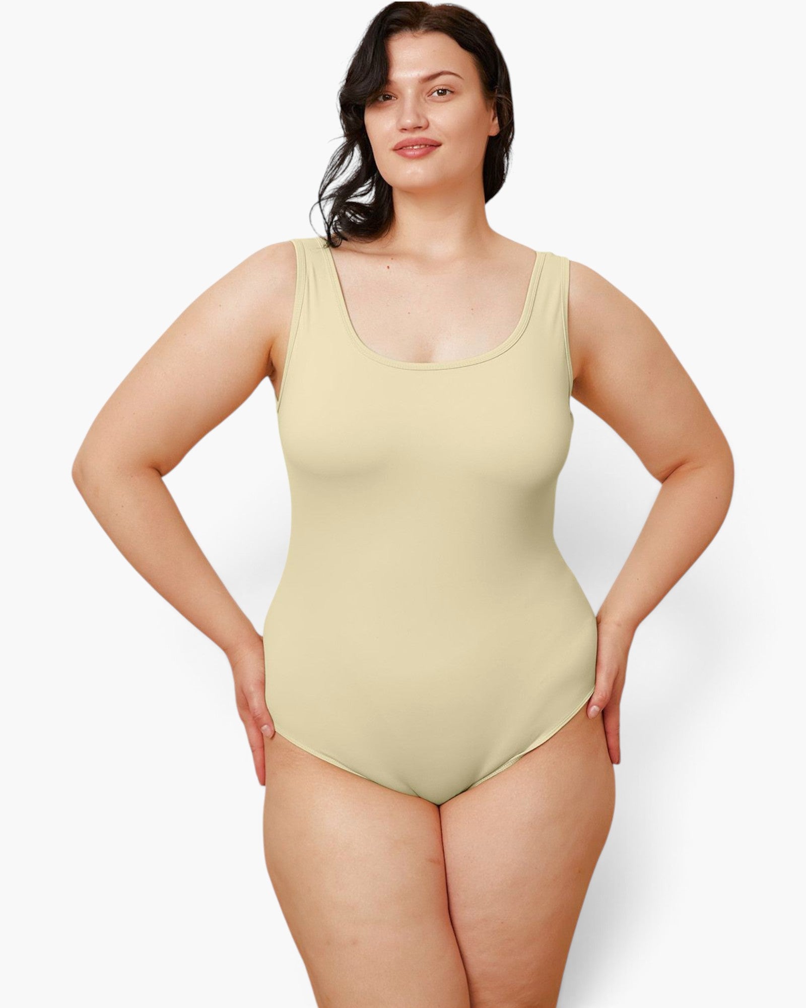 A plus-size model wearing a light beige scoop-neck one-piece swimsuit, standing against a plain light gray studio background.