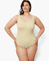 A plus-size model wearing a light beige scoop-neck one-piece swimsuit, standing against a plain light gray studio background.