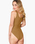 A woman modeling a sleeveless, high-cut one-piece swimsuit in a warm camel brown color, shown from a three-quarters back angle against a light gray studio background.