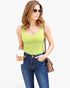 A woman wearing a bright lime green scoop-neck tank top and blue jeans, holding a coffee cup and carrying a brown shoulder bag against a plain white background.