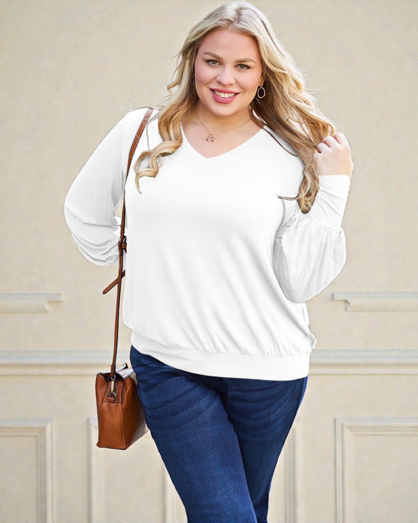 Woman wearing a white long-sleeve top and blue jeans, holding a brown leather handbag against a beige wall.