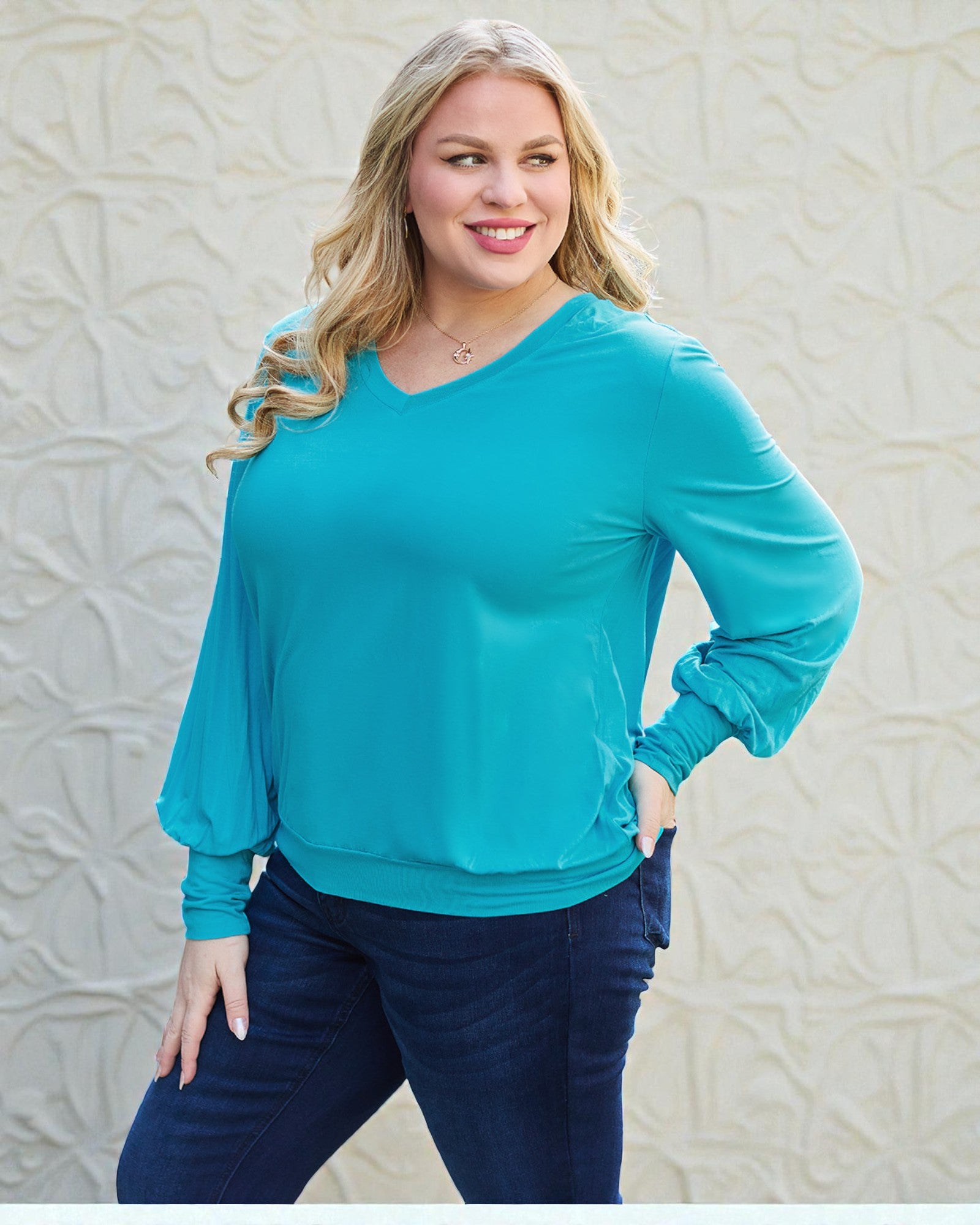 Woman wearing a turquoise sweater and blue pants against a textured beige wall.