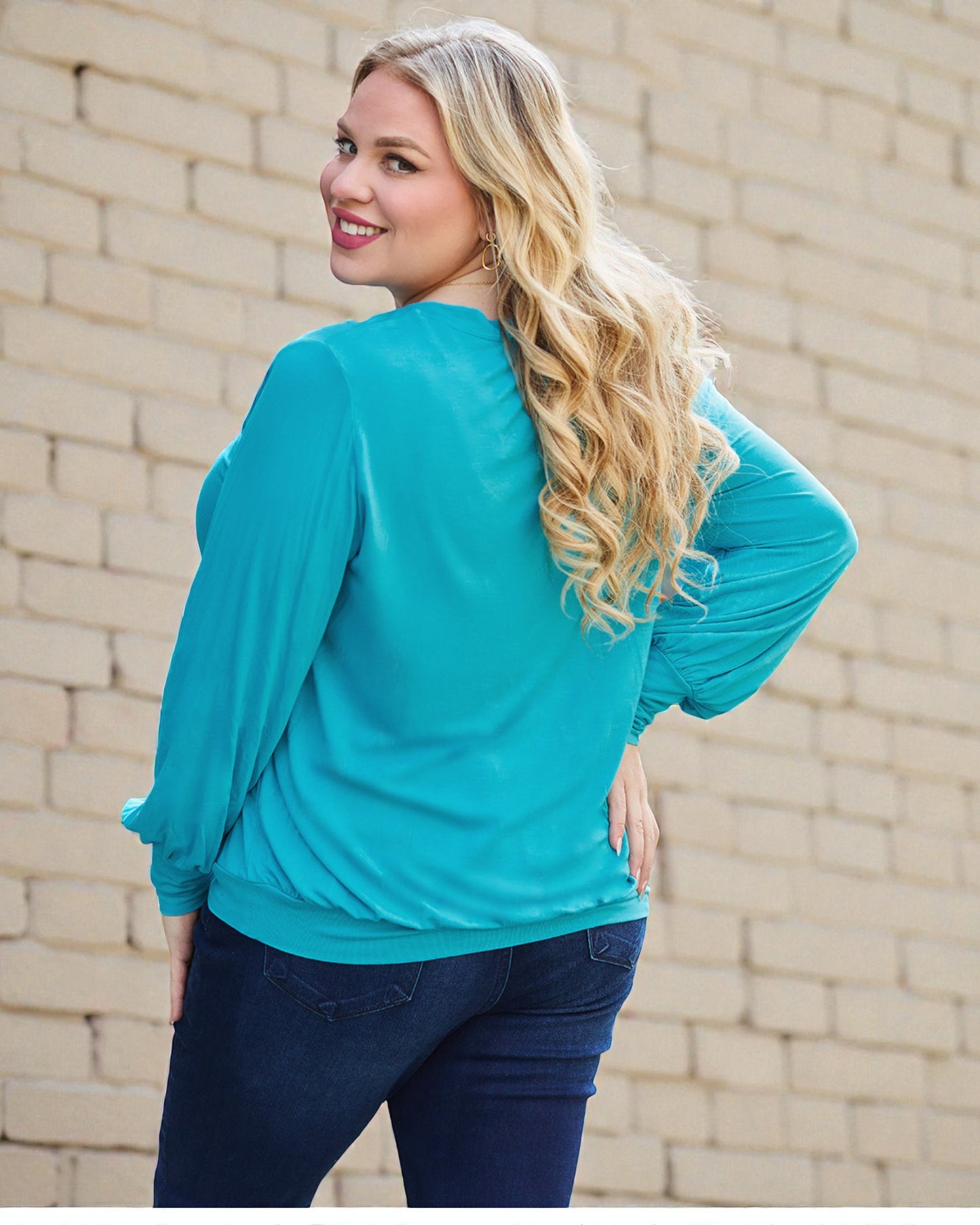 Woman wearing a turquoise long-sleeve top and blue jeans against a beige brick wall.