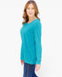 A woman modeling a bright turquoise long-sleeve top with a V-neck, paired with dark blue jeans, against a plain white studio background.