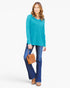 A woman modeling a bright turquoise long-sleeve top with blue jeans and a brown leather handbag, standing against a plain white studio background.