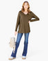 A woman modeling a long-sleeve olive green V-neck top with a chest pocket, paired with blue jeans and holding a light beige backpack, against a plain white background.