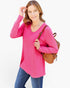 A woman modeling a bright pink long-sleeve V-neck top with blue jeans, carrying a brown woven backpack against a plain white background.