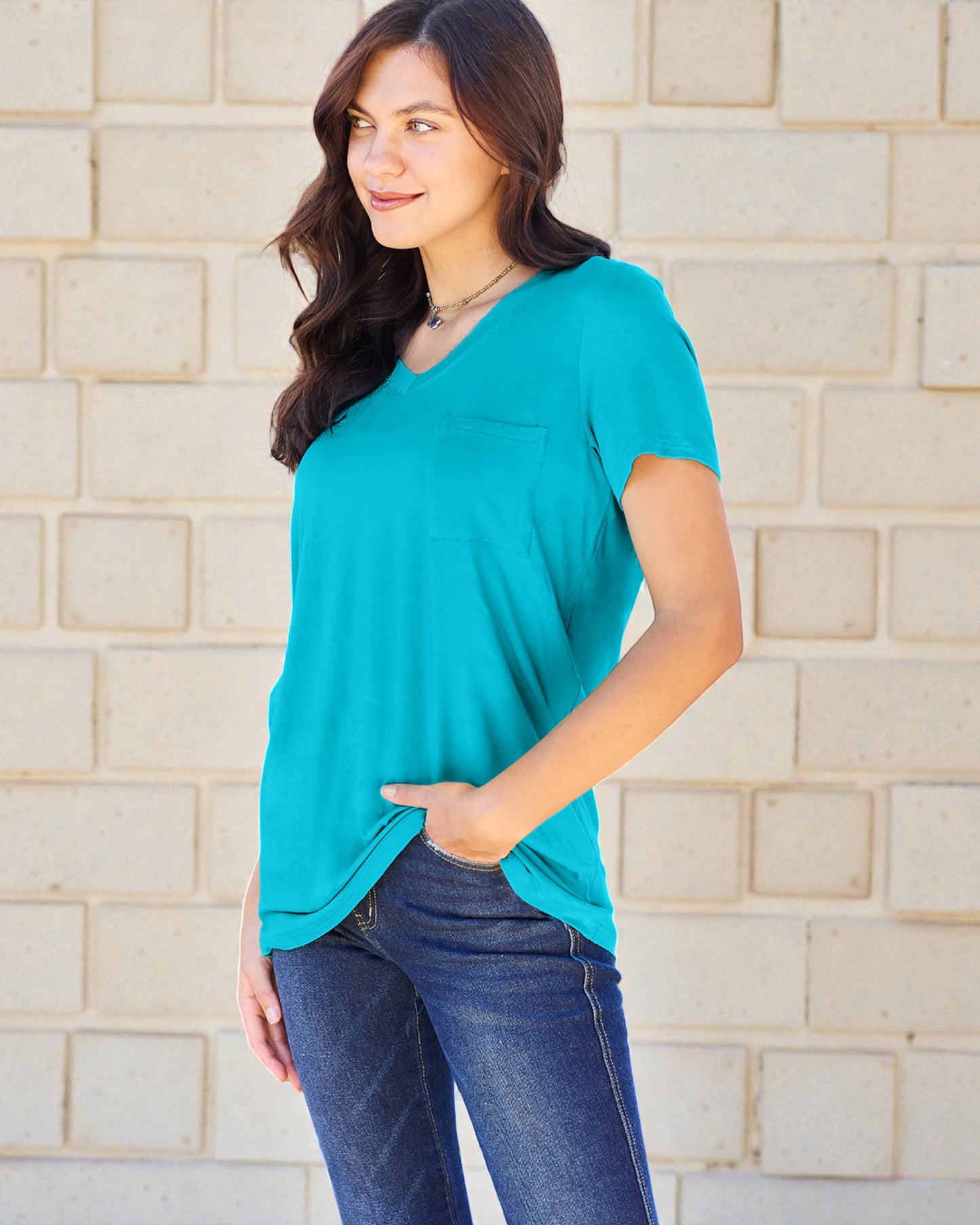 Woman wearing a teal top and blue jeans standing against a beige brick wall.