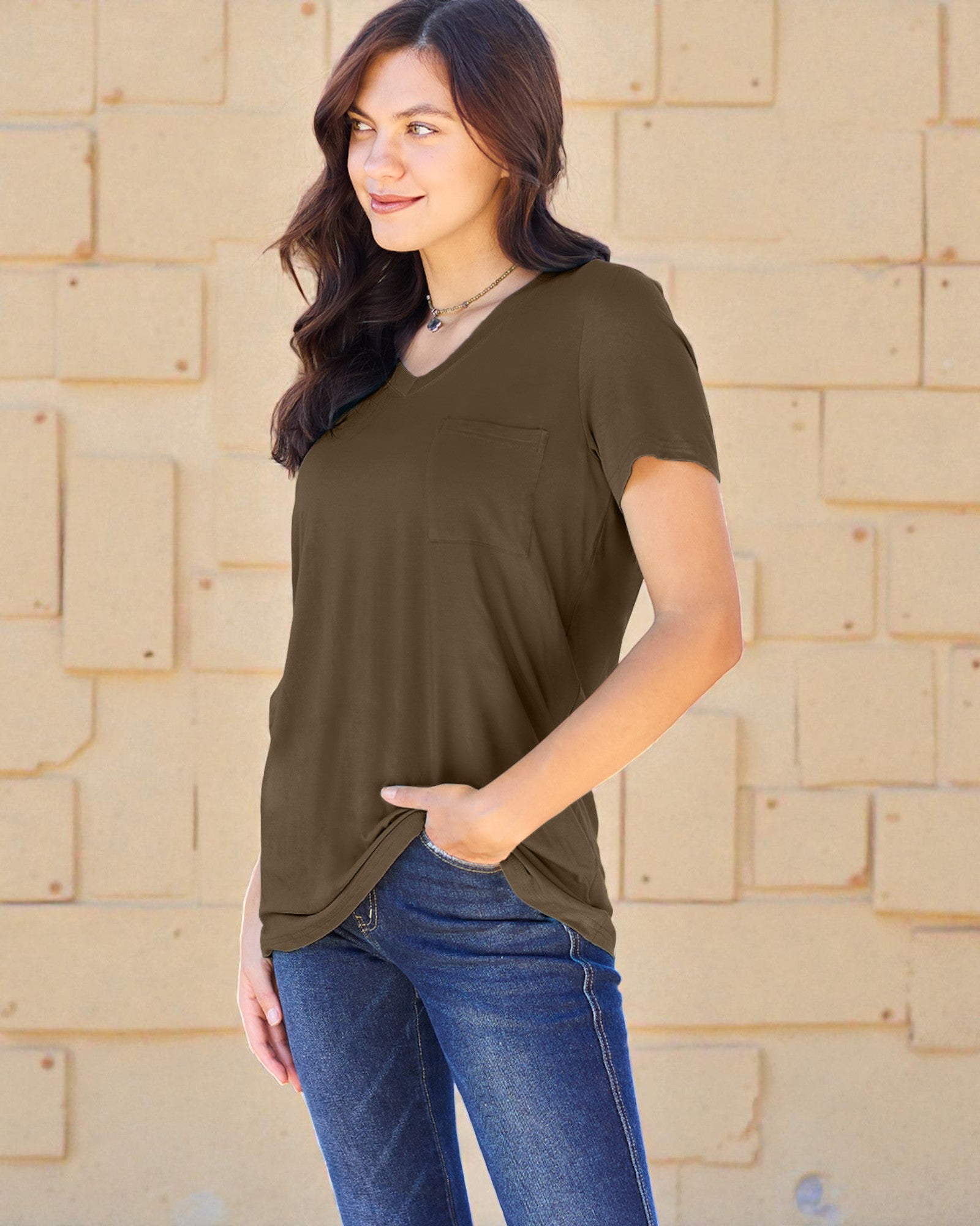 Woman wearing an olive green t-shirt and blue jeans against a beige brick wall.