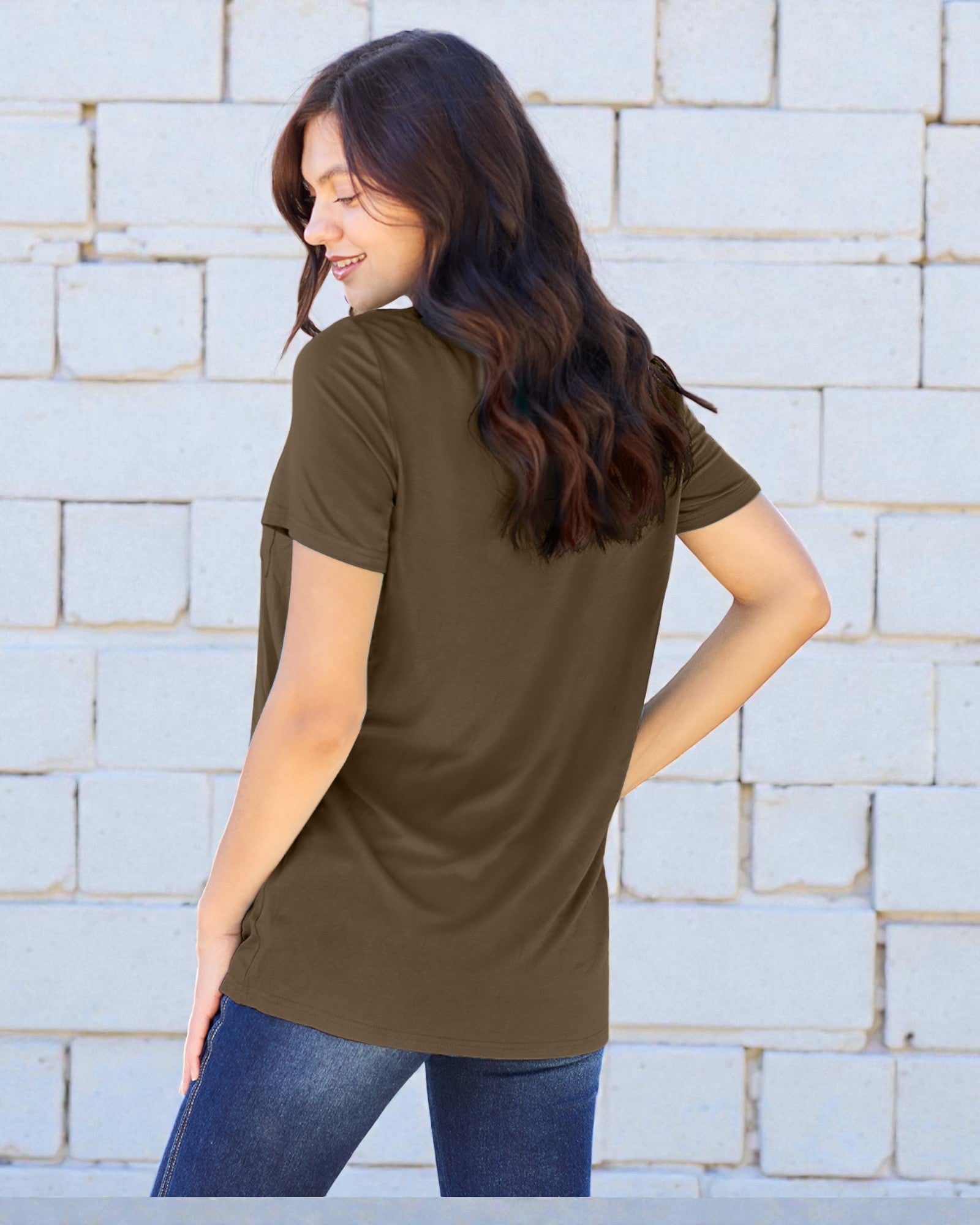 Woman wearing an olive green t-shirt and blue jeans against a light brick wall.