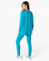 A woman modeling a matching turquoise blue lounge set consisting of a long-sleeve top and leggings, shown from a rear three-quarter angle against a plain white studio background.