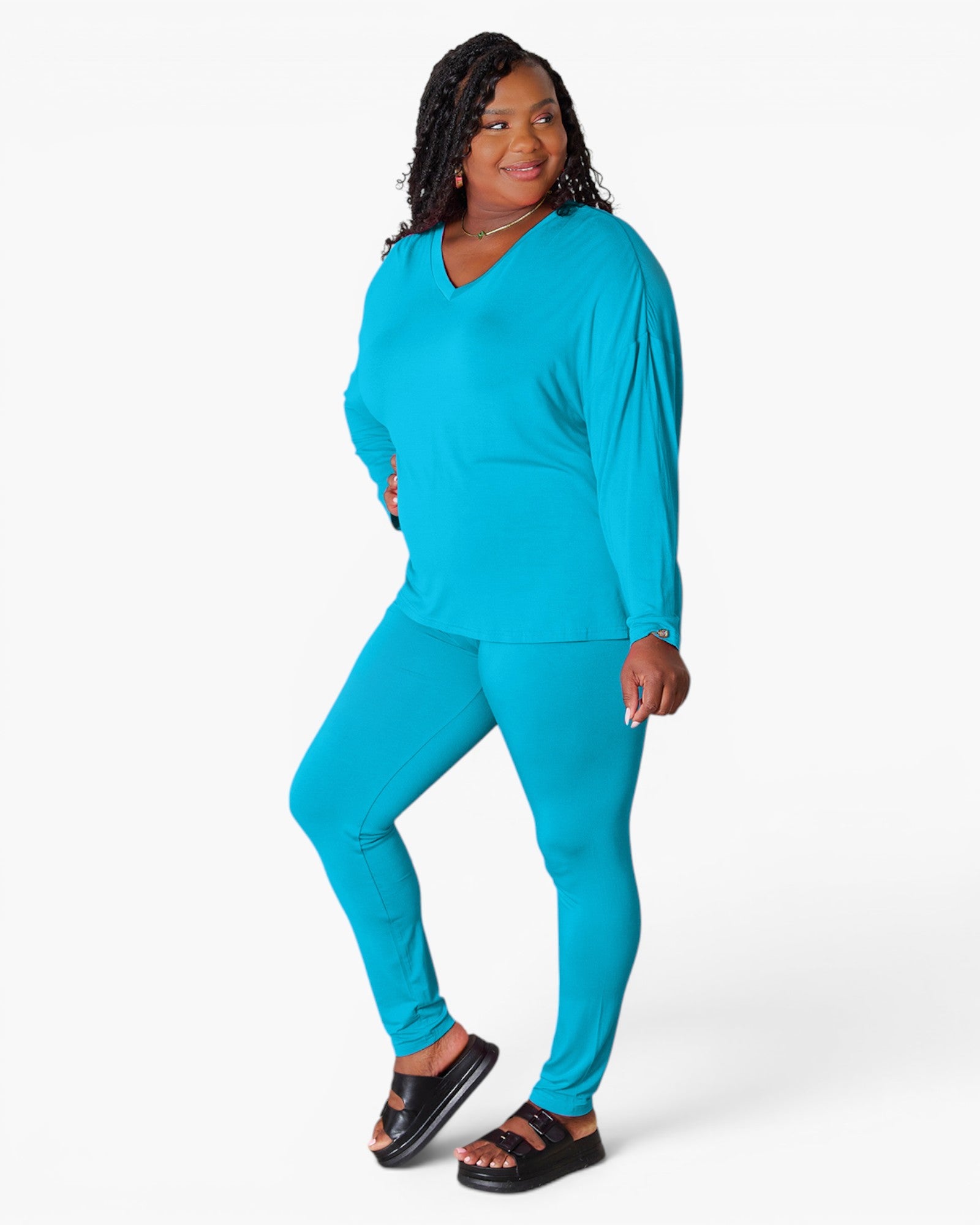 A woman modeling a matching turquoise blue lounge set consisting of a long-sleeve V-neck top and leggings, paired with black slide sandals, against a plain white studio background.