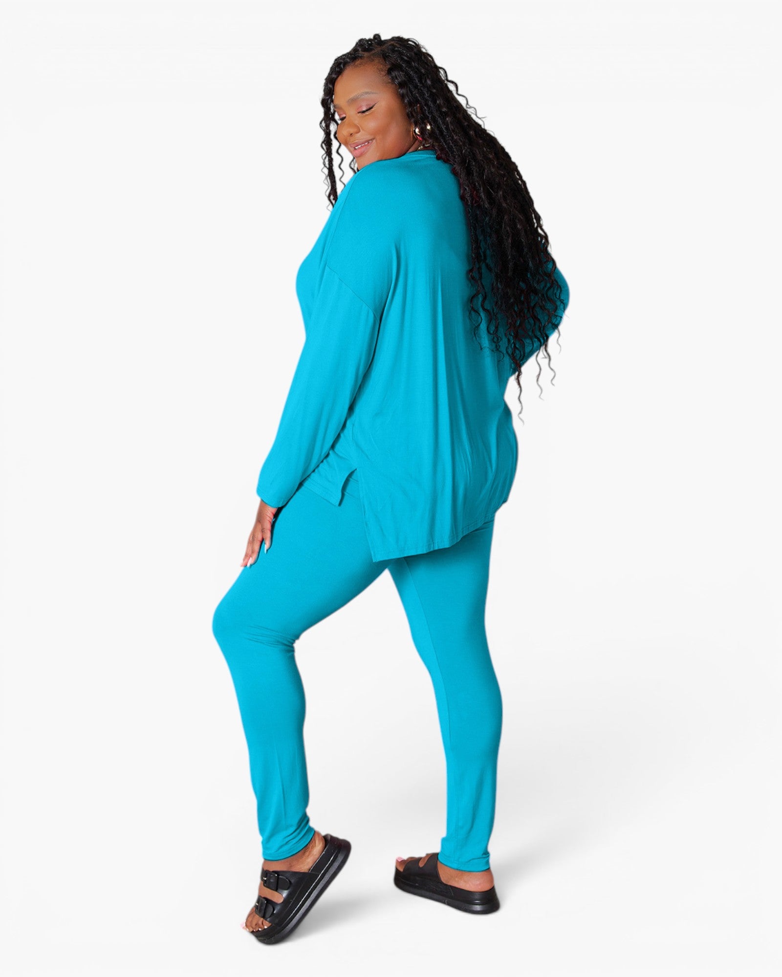 A woman modeling a matching turquoise blue lounge set consisting of a long-sleeve top and leggings, paired with black slide sandals, against a plain white studio background.