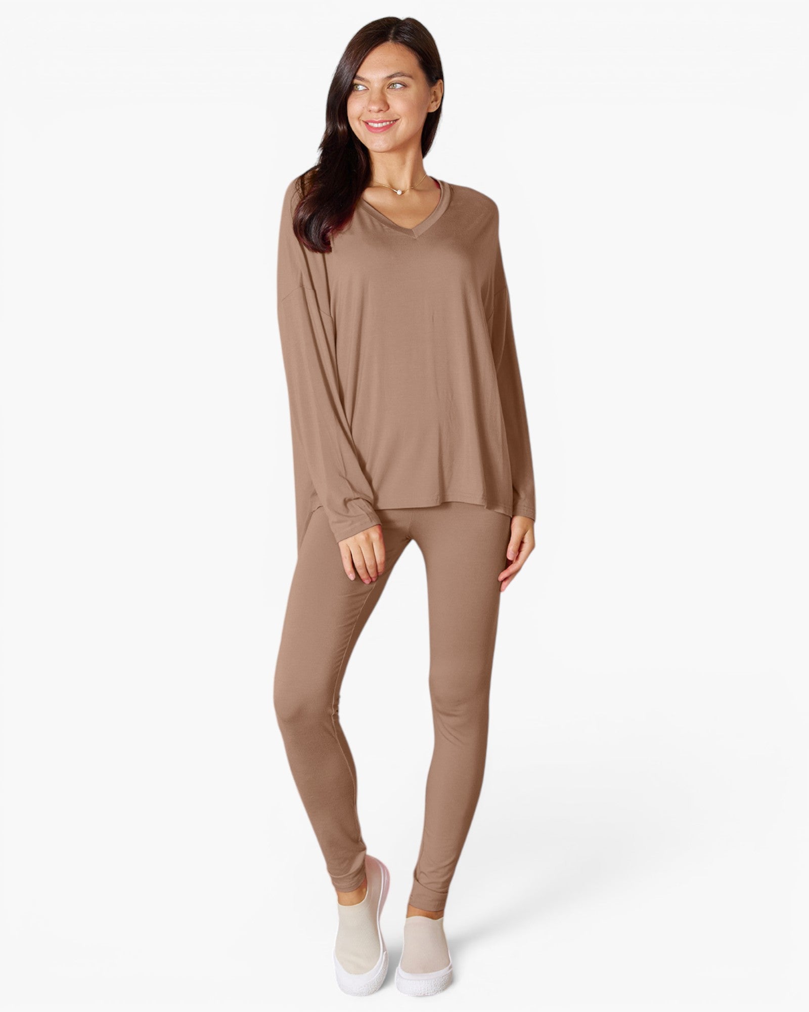 A woman modeling a matching two-piece loungewear set in a solid taupe-brown color, featuring a long-sleeve V-neck top and matching leggings, paired with white slip-on shoes against a plain white studio background.