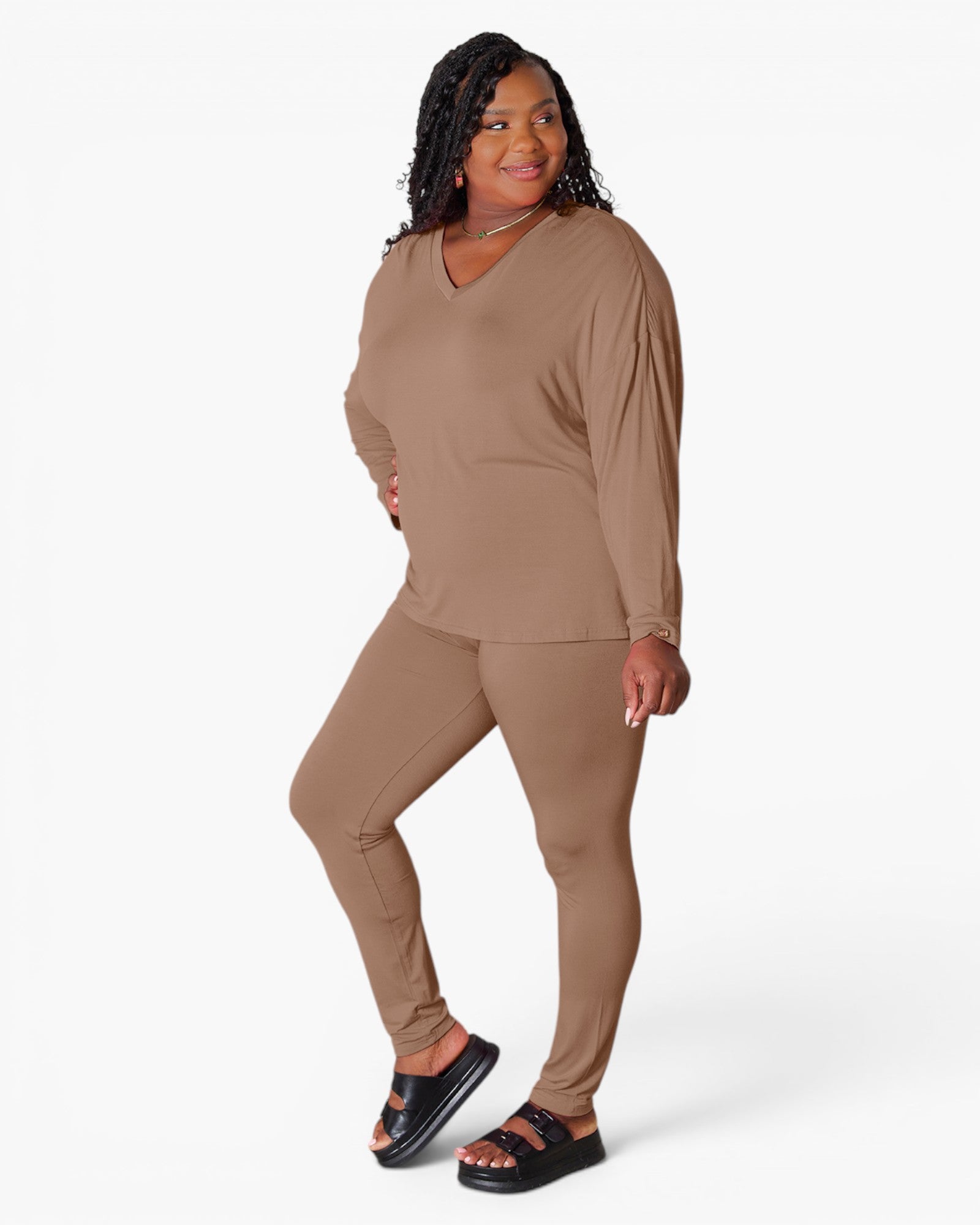 A woman modeling a matching two-piece lounge set in a solid taupe-brown color, featuring a long-sleeve V-neck top and matching leggings, paired with black slide sandals against a plain white studio background.