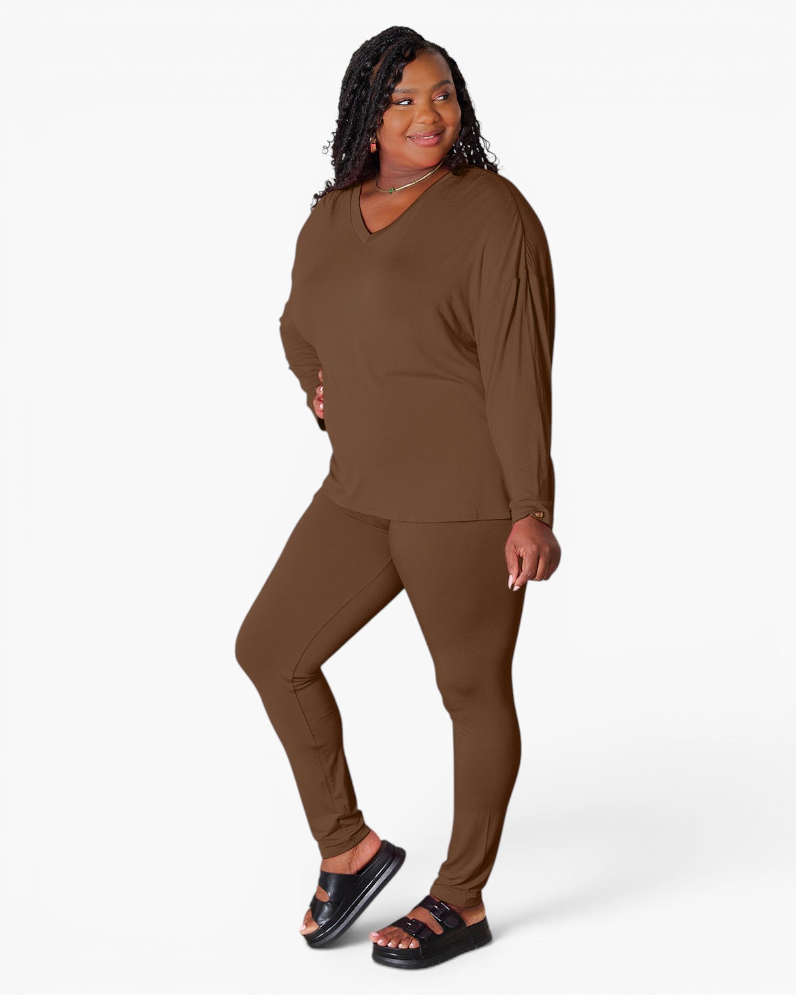 A woman modeling a matching brown lounge set consisting of a long-sleeve V-neck top and high-waisted leggings, paired with black slide sandals, against a plain white studio background.
