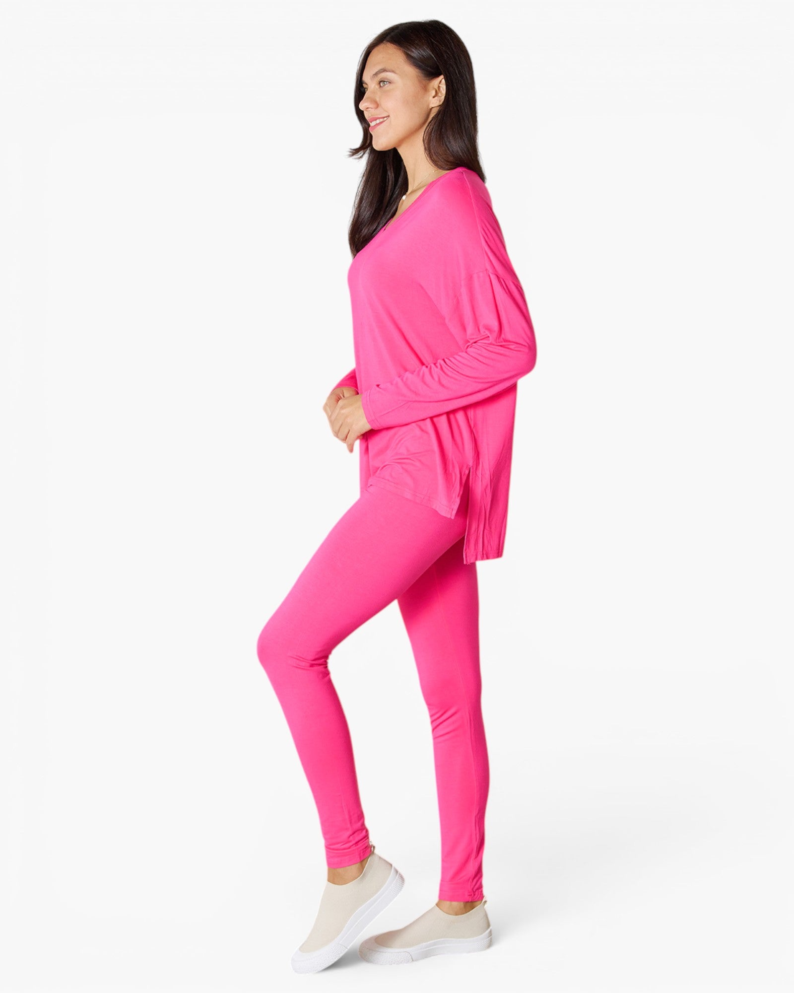 A woman modeling a bright pink activewear set consisting of a long-sleeve V-neck top and matching leggings, paired with light beige slip-on sneakers, against a plain white studio background.