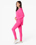 A woman modeling a bright pink activewear set consisting of a long-sleeve V-neck top and matching leggings, paired with light beige slip-on sneakers, against a plain white studio background.