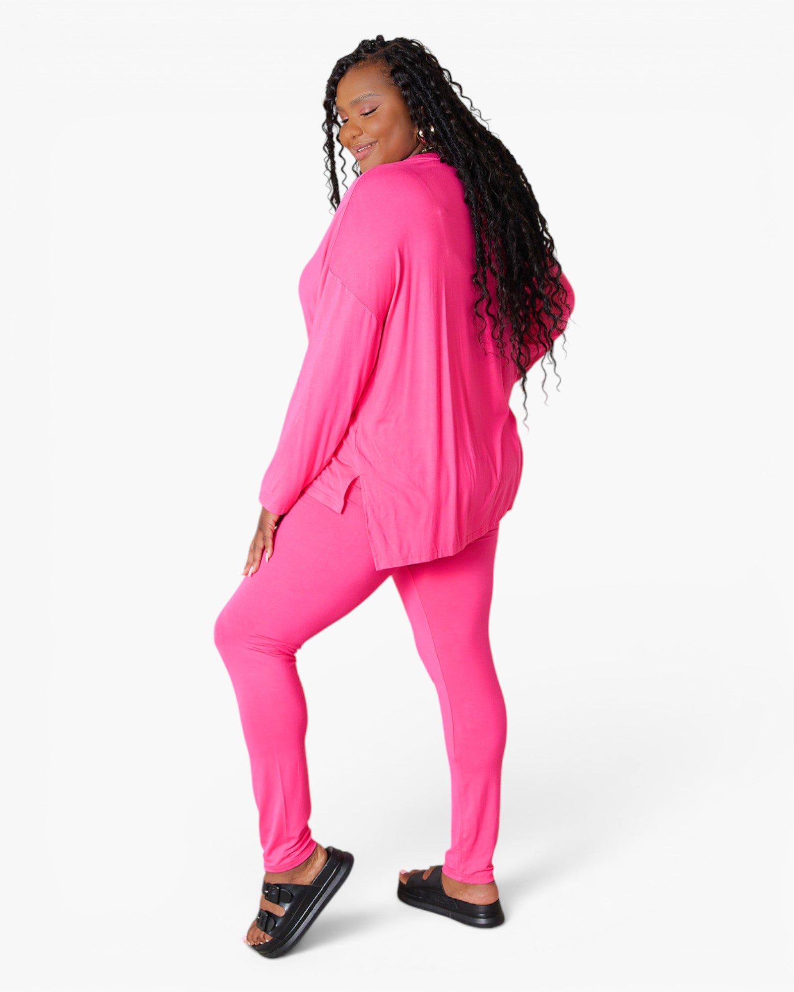 A woman modeling a bright pink lounge set consisting of a long-sleeve top and matching leggings, paired with black slide sandals, against a plain white studio background.