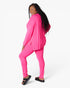 A woman modeling a bright pink lounge set consisting of a long-sleeve top and matching leggings, paired with black slide sandals, against a plain white studio background.