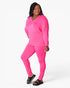 A woman modeling a bright pink matching lounge set consisting of a long-sleeve V-neck top and leggings, paired with black slide sandals, against a plain white studio background.