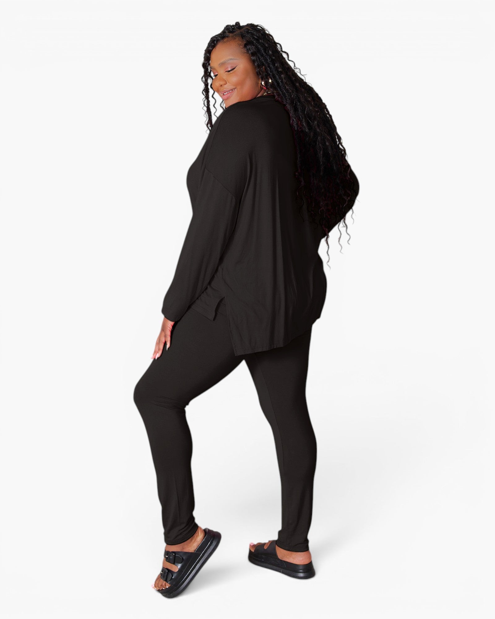 A woman modeling a black two-piece loungewear set consisting of a long-sleeve draped top and matching slim-fit pants, paired with black slide sandals, against a plain white studio background.