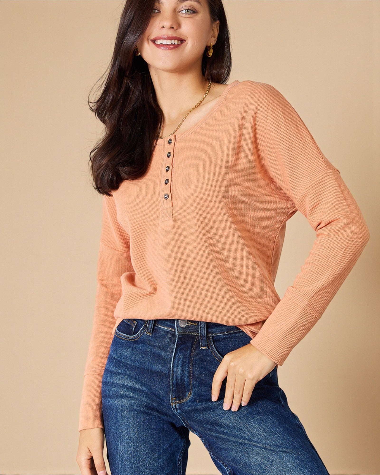 Woman wearing a peach long-sleeve top and blue jeans against a beige background