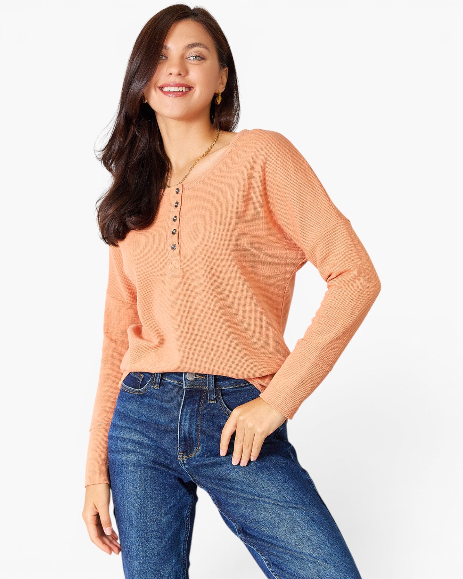 A woman wearing a peach-colored long-sleeve henley top with blue jeans, standing against a plain white studio background.