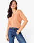 A woman wearing a peach-colored long-sleeve henley top with blue jeans, standing against a plain white studio background.