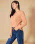 Woman wearing a peach long-sleeve top and blue jeans against a beige background