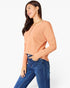 A woman wearing a peach long-sleeve knit top with a decorative button detail at the neckline, paired with blue jeans, photographed against a clean white studio background.