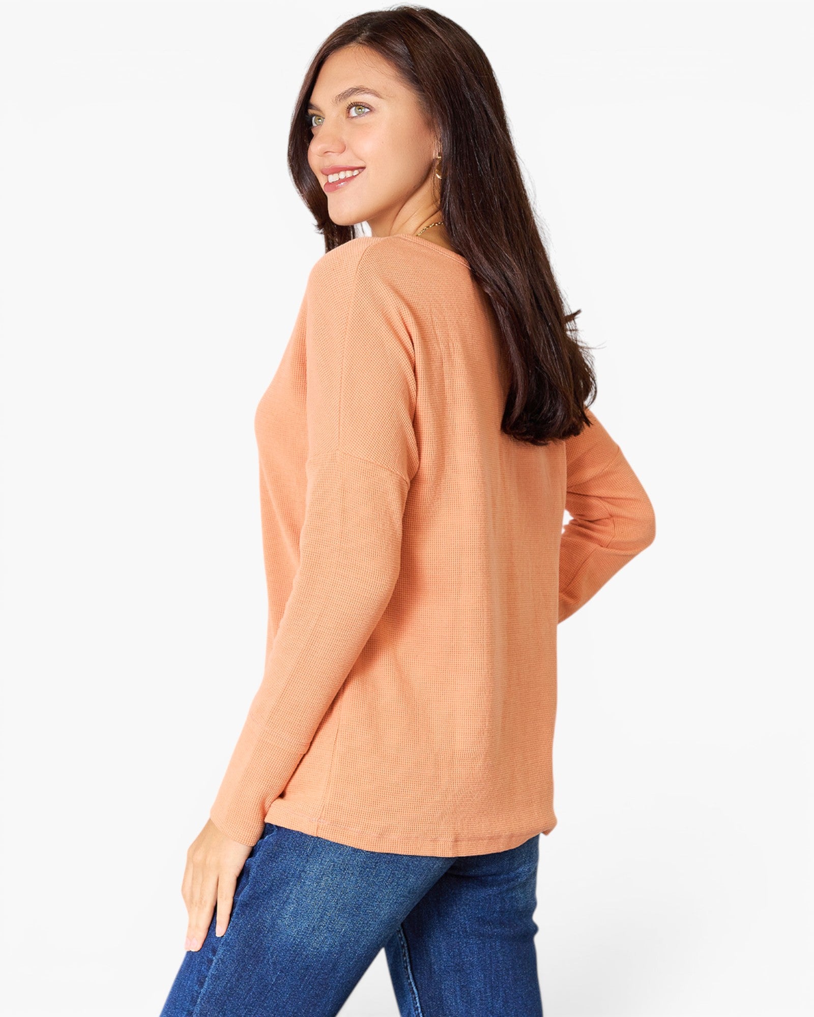 A woman wearing a peach-colored long-sleeve top and blue jeans, photographed from the back against a plain white studio background.
