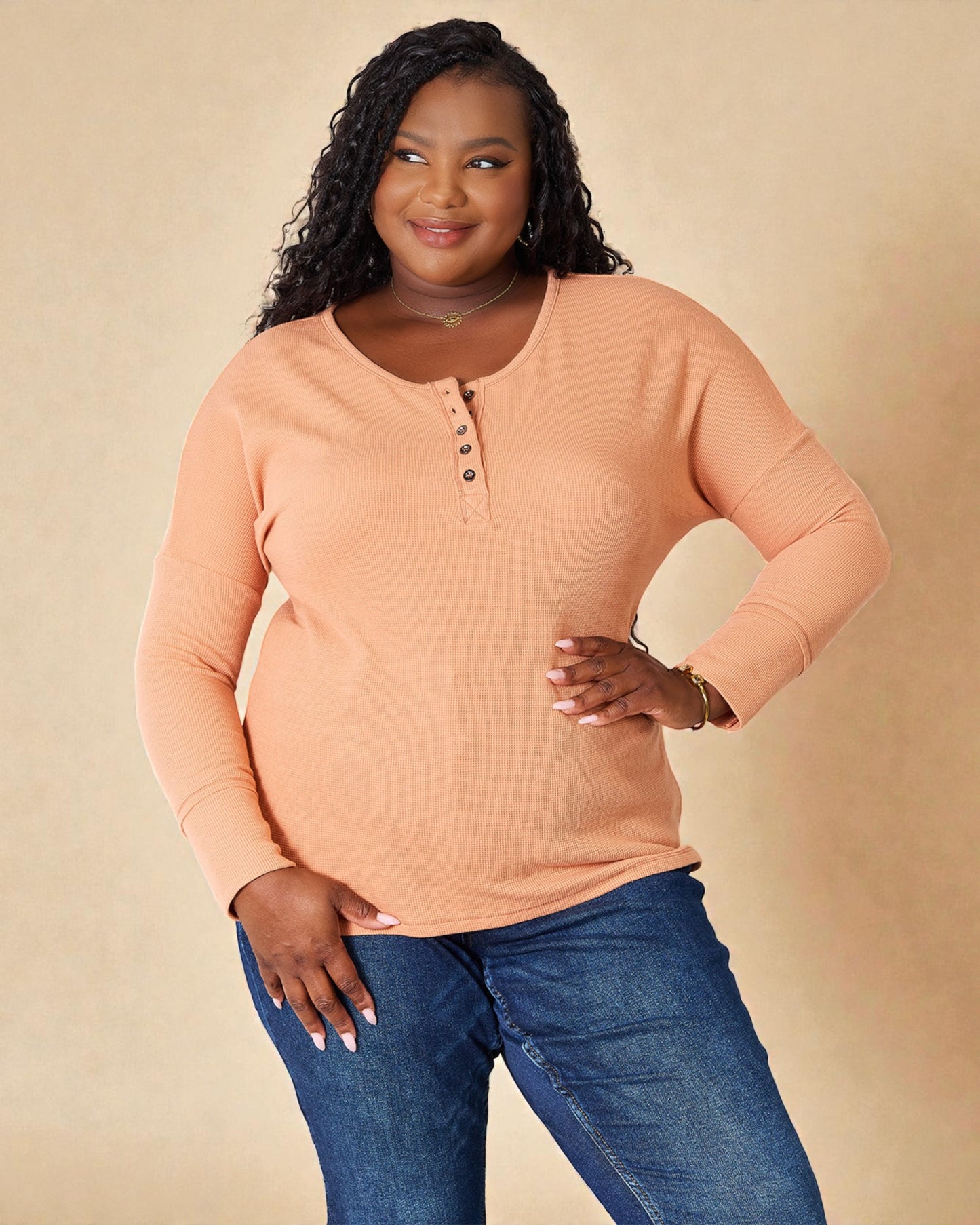 Woman wearing a peach long-sleeve henley shirt and blue jeans against a beige background
