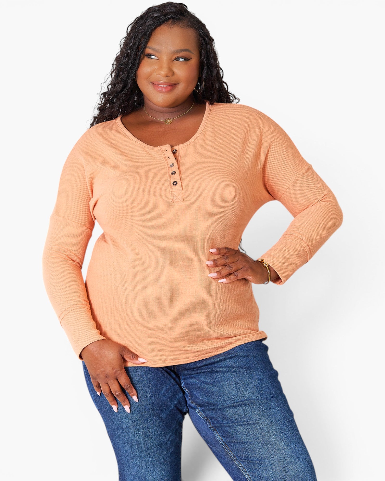 A woman modeling a peach-colored long-sleeve henley top with blue jeans against a plain white studio background.