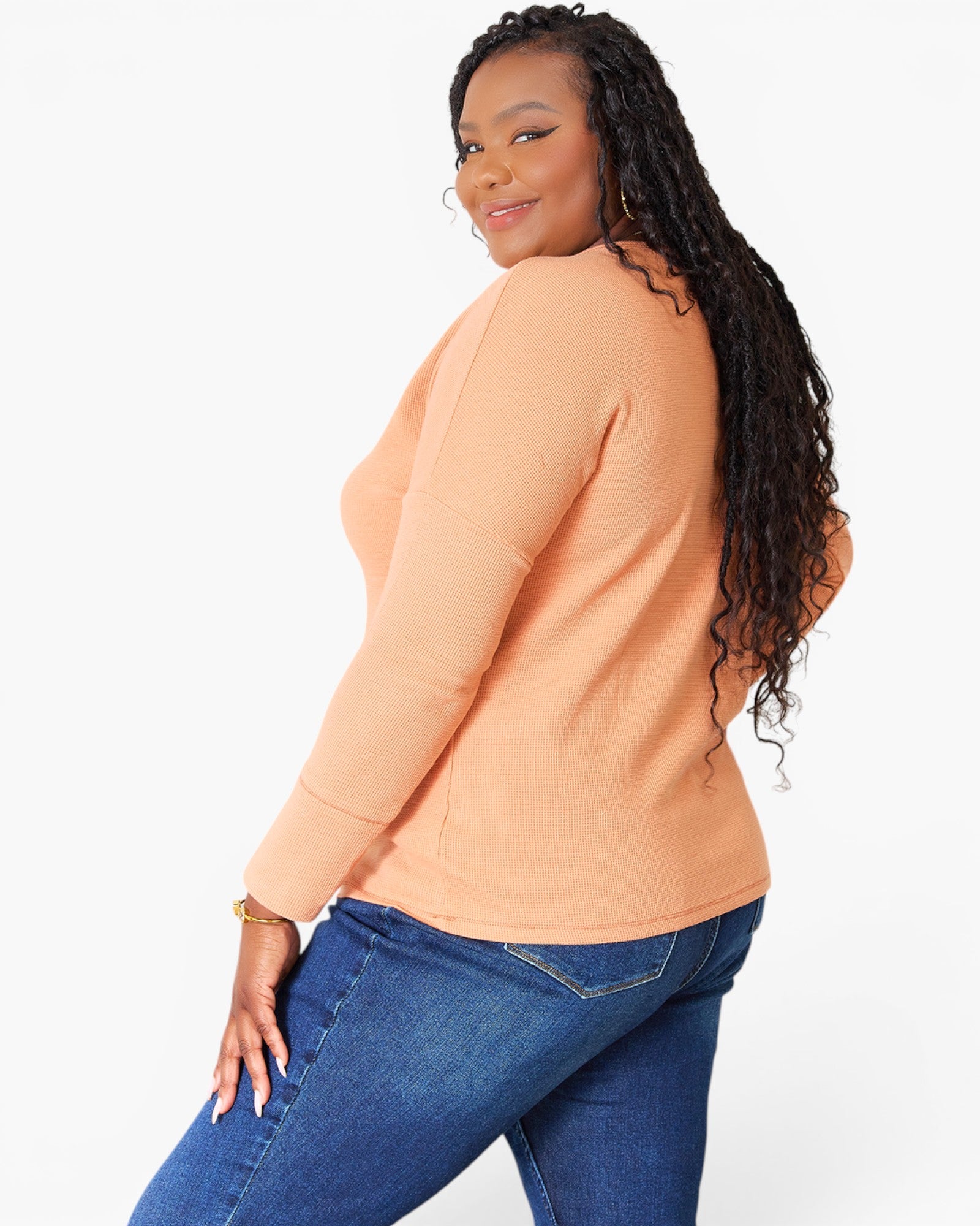 A woman with long dark curly hair is shown from a side-back angle wearing a peach-colored long-sleeve knit top and blue jeans against a plain white studio background.