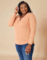 Woman wearing a peach long-sleeve top and blue jeans against a beige background