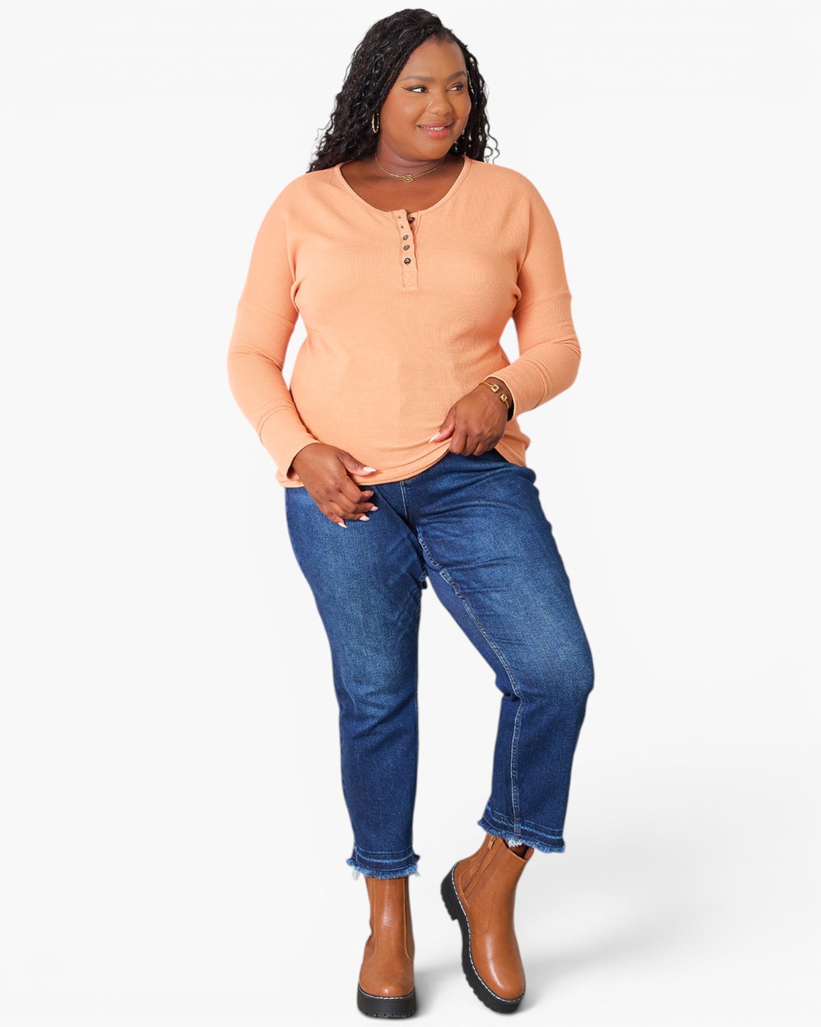 A woman modeling a peach long-sleeve henley top with blue jeans and brown ankle boots, standing against a light gray studio background.