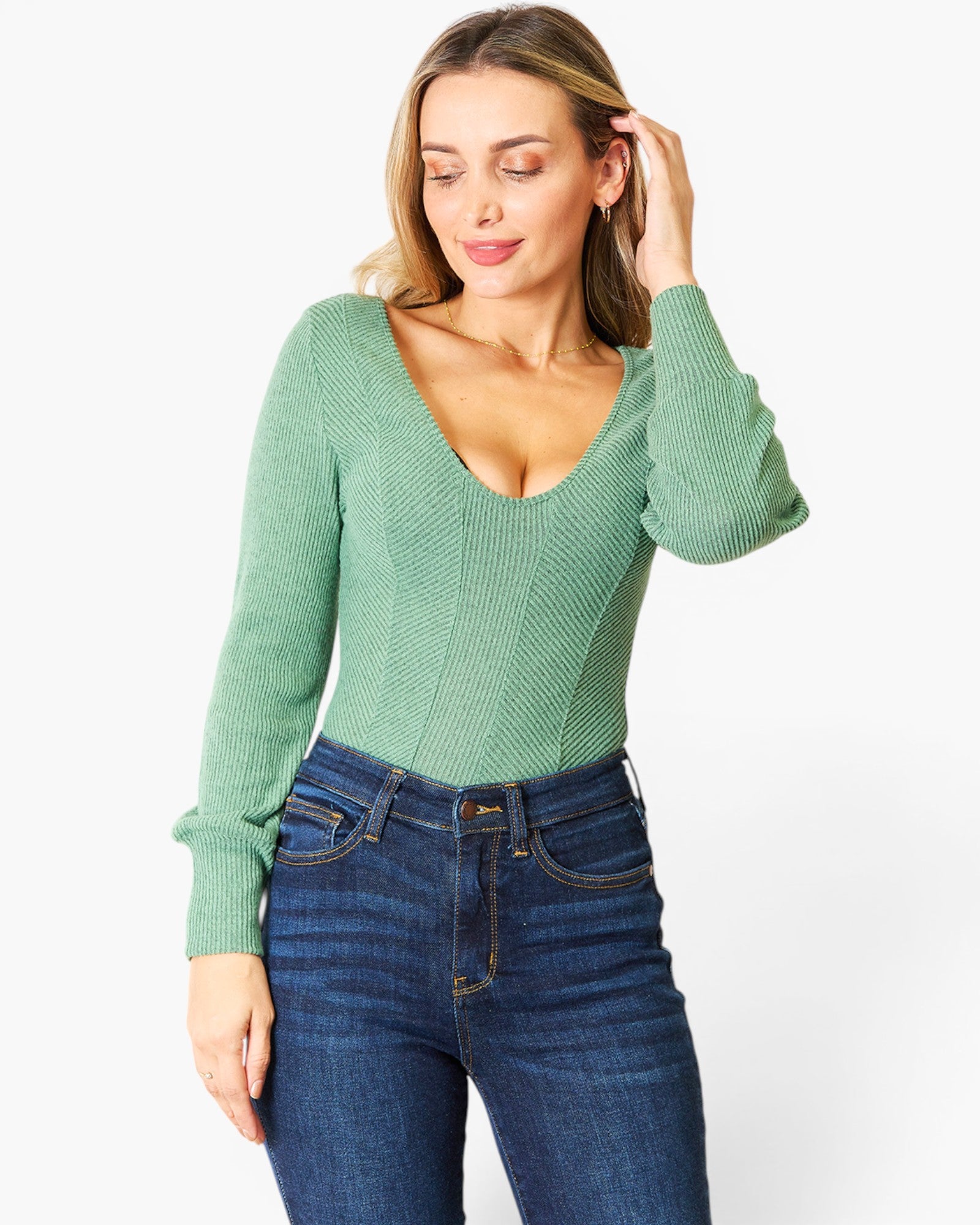 A woman wearing a sage green ribbed knit long-sleeve top with a deep V-neck and dark blue jeans, posing against a plain white studio background.