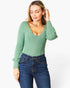 A woman wearing a sage green ribbed knit long-sleeve top with a deep V-neck and dark blue jeans, posing against a plain white studio background.
