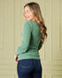 Woman wearing a green sweater and blue jeans against a beige paneled wall.