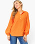 A woman wearing a bright orange long-sleeve henley-style top with a chest pocket and three front buttons, paired with blue jeans, against a plain light background.