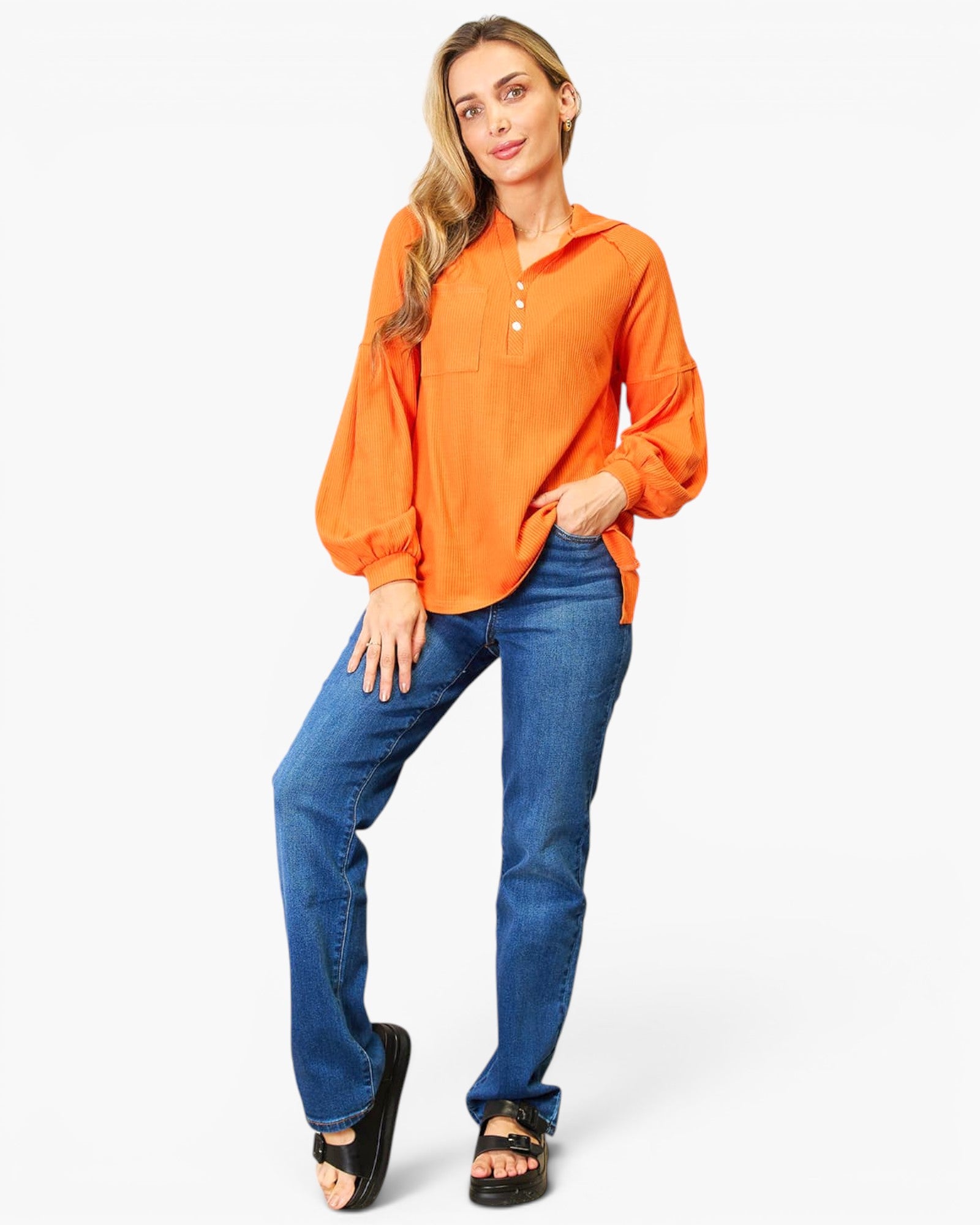 A woman modeling a bright orange long-sleeve button-up shirt with blue jeans and black sandals, standing against a plain white studio background.