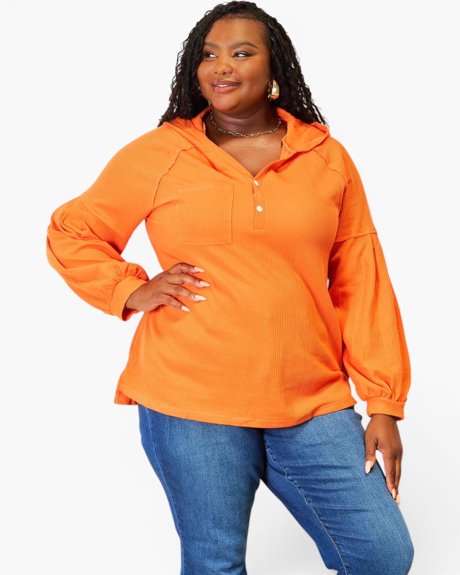 A woman modeling a bright orange long-sleeve pullover top with a small button placket and hood, paired with blue jeans, against a plain white studio background.