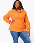 A woman modeling a bright orange long-sleeve pullover top with a small button placket and hood, paired with blue jeans, against a plain white studio background.