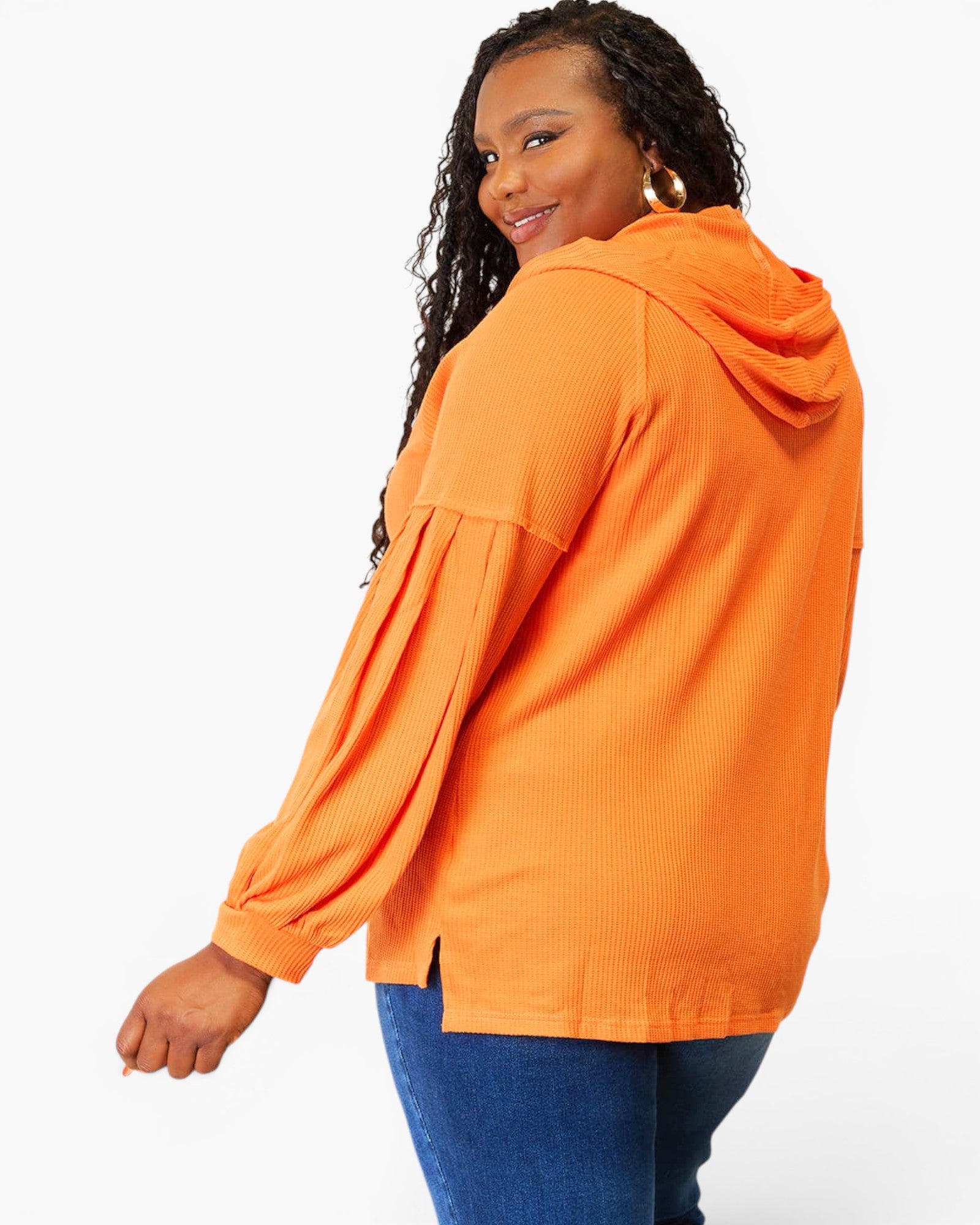 A woman wearing a bright orange hooded long-sleeve top with pleated sleeves and blue jeans, photographed against a clean white studio background.
