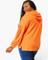 A woman wearing a bright orange hooded long-sleeve top with pleated sleeves and blue jeans, photographed against a clean white studio background.