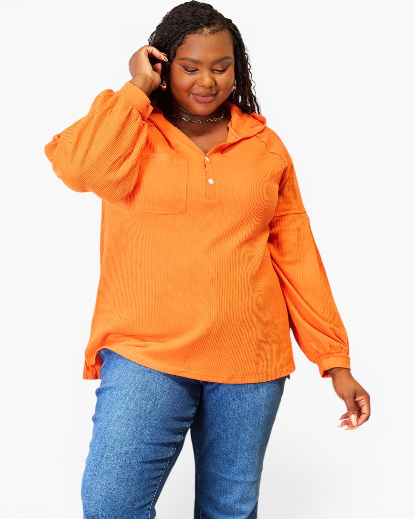 A woman modeling a bright orange long-sleeve pullover top with a small button placket and chest pocket, paired with blue jeans, against a plain light gray background.