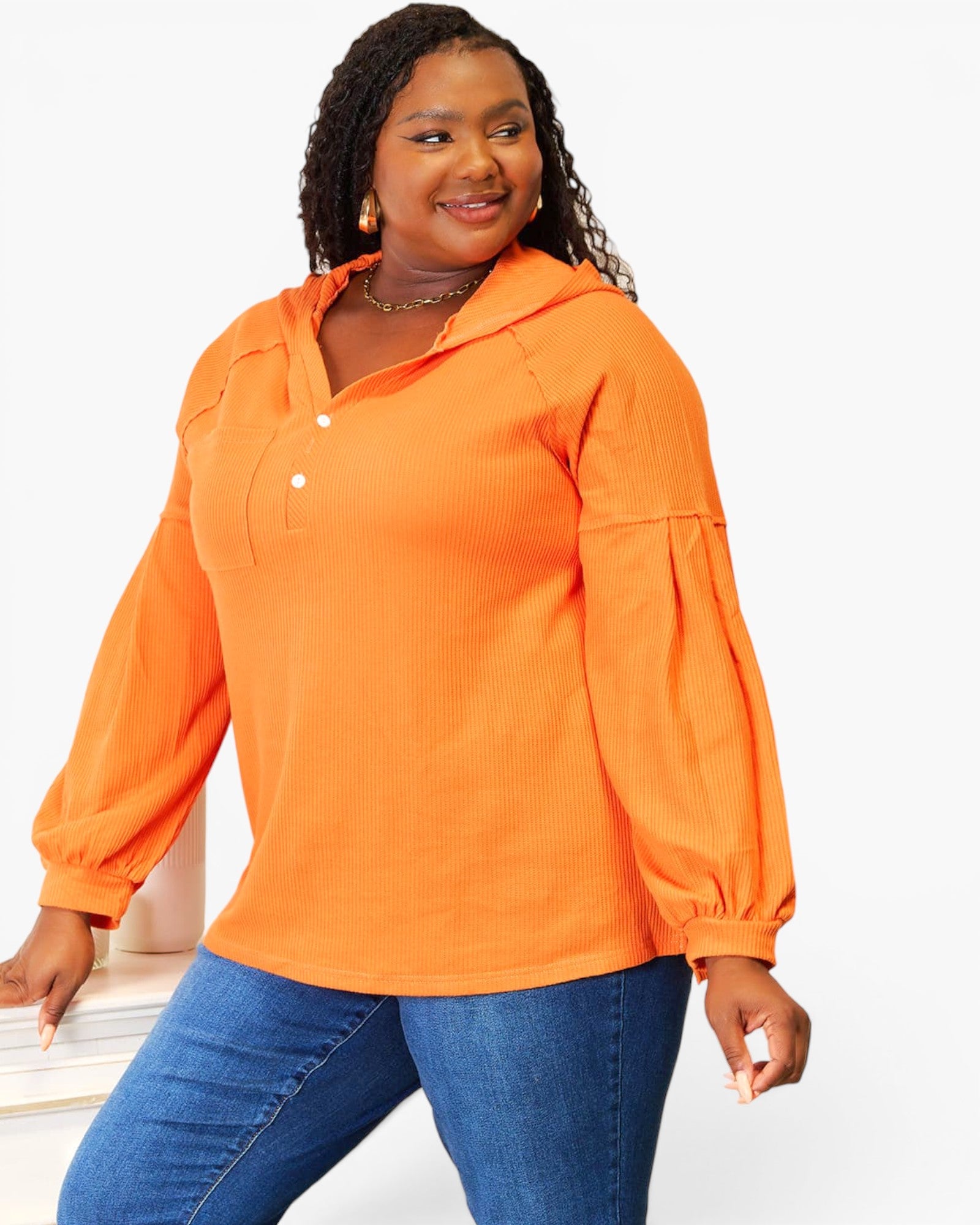 A woman modeling a bright orange long-sleeve pullover top with a buttoned collar and puff sleeves, paired with blue jeans, against a plain white studio background.